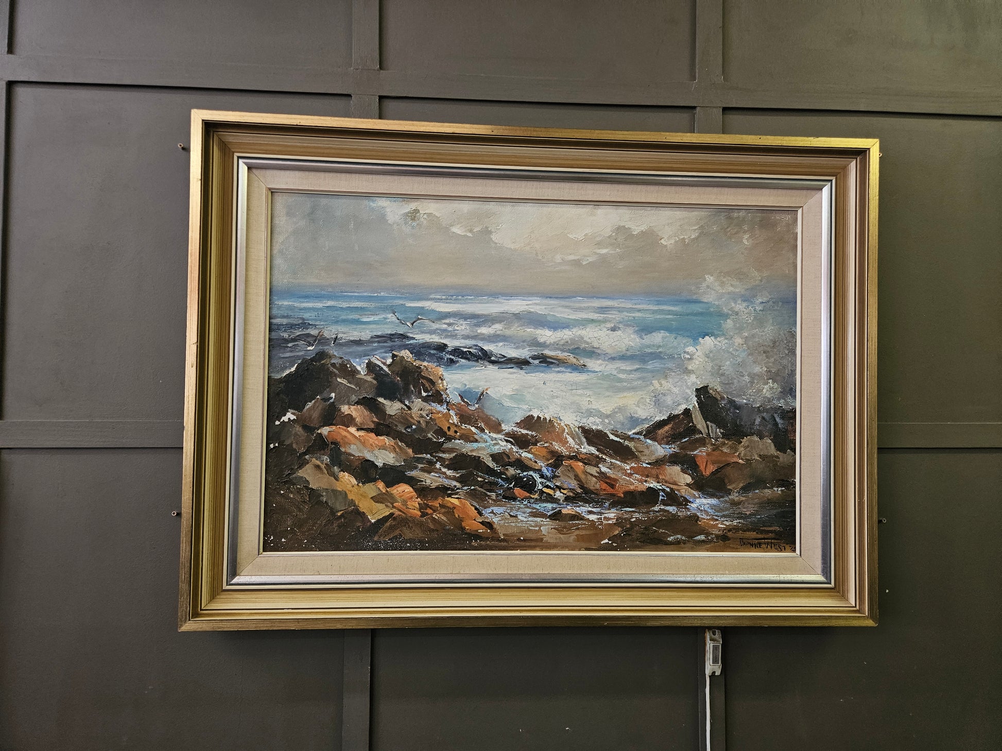 Framed painting of a coastal scene with rocks and water on a gray wall.